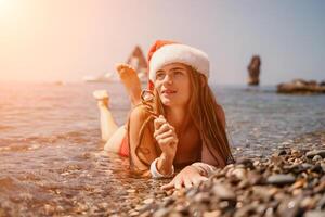 Woman travel sea. Happy tourist enjoy taking picture on the beach for memories. Woman traveler in Santa hat looks at camera on the sea bay, sharing travel adventure journey photo