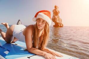 mujer mar sorber. cerca arriba retrato de contento joven caucásico mujer con largo pelo en Papa Noel sombrero mirando a cámara y sonriente. linda mujer retrato en un blanco bikini posando en cenar tablero en el mar foto