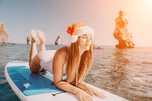 mujer mar sorber. cerca arriba retrato de contento joven caucásico mujer con largo pelo en Papa Noel sombrero mirando a cámara y sonriente. linda mujer retrato en un blanco bikini posando en cenar tablero en el mar foto