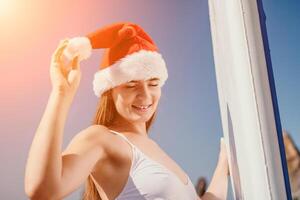 mujer mar sorber. cerca arriba retrato de contento joven caucásico mujer con largo pelo en Papa Noel sombrero mirando a cámara y sonriente. linda mujer retrato en un blanco bikini posando en cenar tablero en el mar foto