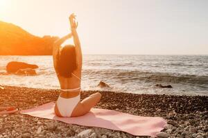 Woman sea yoga. Happy woman in white swimsuit and boho style braclets practicing outdoors on yoga mat by sea on sunset. Women yoga fitness routine. Healthy lifestyle, harmony and meditation photo