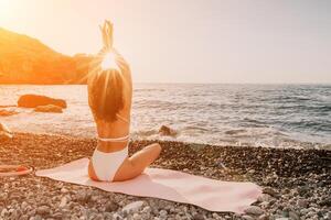 Woman sea yoga. Happy woman in white swimsuit and boho style braclets practicing outdoors on yoga mat by sea on sunset. Women yoga fitness routine. Healthy lifestyle, harmony and meditation photo