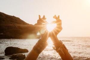 Woman sea yoga. Happy woman in a white swimsuit and boho-style bracelets making a heart shape with her hands while practicing yoga on a mat by sea at sunset. Healthy lifestyle, harmony and meditation photo