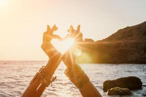 Woman sea yoga. Happy woman in a white swimsuit and boho-style bracelets making a heart shape with her hands while practicing yoga on a mat by sea at sunset. Healthy lifestyle, harmony and meditation photo