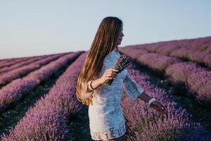 mujer lavanda campo. contento despreocupado mujer en un blanco vestir caminando en un lavanda campo y oliendo un lavanda ramo de flores en puesta de sol. ideal para calentar y inspirador conceptos en pasión de viajar y viajar. foto