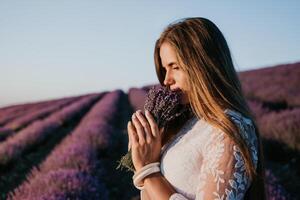 Woman lavender field. Happy carefree woman in a white dress walking in a lavender field and smelling a lavender bouquet on sunset. Ideal for warm and inspirational concepts in wanderlust and travel. photo