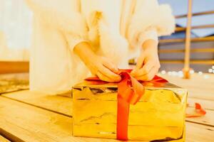 A woman in a white dress is holding a gold box with a red ribbon. She is wearing a crown on her head. The woman appears to be opening the gift box. photo