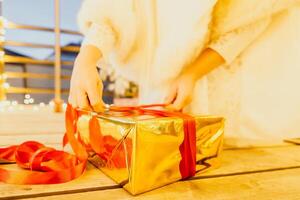 A woman in a white dress is holding a gold box with a red ribbon. She is wearing a crown on her head. The woman appears to be opening the gift box. photo