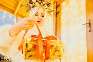 un mujer en un blanco vestir es participación un oro caja con un rojo cinta. ella es vistiendo un corona en su cabeza. el mujer aparece a ser apertura el regalo caja. foto