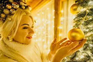 A blonde woman in white dress and a crown of gold ornaments decorate Christmas tree with gold ornaments and lights. The tree is decorated with gold balls and is lit up with lights. photo