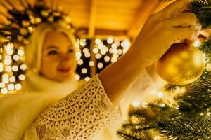 A blonde woman in white dress and a crown of gold ornaments decorate Christmas tree with gold ornaments and lights. The tree is decorated with gold balls and is lit up with lights. photo