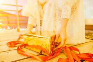 A woman in a white dress is holding a gold box with a red ribbon. She is wearing a crown on her head. The woman appears to be opening the gift box. photo