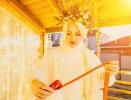 A woman in a white dress is holding a gold box with a red ribbon. She is wearing a crown on her head. The woman appears to be opening the gift box. photo