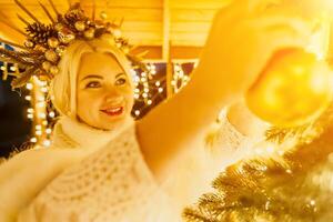 un rubia mujer en blanco vestir y un corona de oro adornos Decorar Navidad árbol con oro adornos y luces. el árbol es decorado con oro pelotas y es iluminado arriba con luces. foto