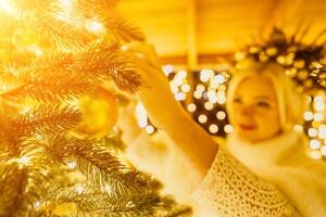 A blonde woman in white dress and a crown of gold ornaments decorate Christmas tree with gold ornaments and lights. The tree is decorated with gold balls and is lit up with lights. photo