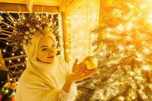 un rubia mujer en blanco vestir y un corona de oro adornos Decorar Navidad árbol con oro adornos y luces. el árbol es decorado con oro pelotas y es iluminado arriba con luces. foto