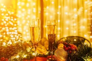 Two champagne glasses filled with champagne are placed on a table near to Christmas tree. The glasses are surrounded by lights, creating a festive atmosphere. photo