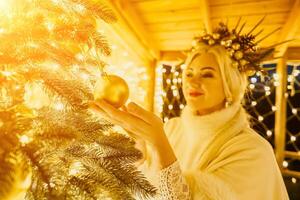 un rubia mujer en blanco vestir y un corona de oro adornos Decorar Navidad árbol con oro adornos y luces. el árbol es decorado con oro pelotas y es iluminado arriba con luces. foto