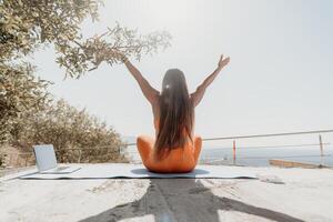 Fitness woman sea. Happy middle aged woman in orange sportswear exercises morning outdoors on yoga mat with laptop in park over ocean beach. Female fitness pilates yoga routine. Healthy lifestyle. photo