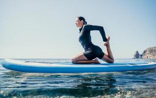 Woman sup yoga. Happy sporty woman practising yoga pilates on paddle sup surfboard. Female stretching doing workout on sea water. Modern individual female hipster outdoor summer sport activity. photo