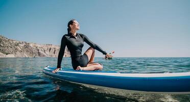 Woman sup yoga. Happy sporty woman practising yoga pilates on paddle sup surfboard. Female stretching doing workout on sea water. Modern individual female hipster outdoor summer sport activity. photo