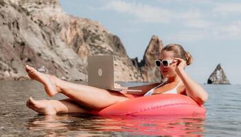 mujer persona de libre dedicación trabajos en ordenador portátil nadando en mar en rosado inflable anillo. contento turista en Gafas de sol flotante en inflable rosquilla y trabajando en ordenador portátil computadora en calma océano. remoto trabajando en cualquier sitio foto