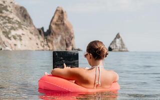 Woman freelancer works on laptop swimming in sea on pink inflatable ring. Happy tourist in sunglasses floating on inflatable donut and working on laptop computer in calm ocean. Remote working anywhere photo