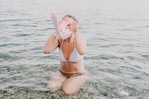 Young happy woman in white bikini and wearing pink mask gets ready for sea snorkeling. Positive smiling woman relaxing and enjoying water activities with family summer travel holidays vacation on sea. photo