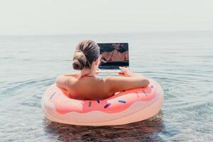 Woman freelancer works on laptop swimming in sea on pink inflatable ring. Happy tourist floating on inflatable donut and working on laptop computer in calm ocean. Freelance, remote working anywhere photo