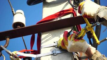 Skilled electrician in helmet fixes wires standing on ladder near high pole against blue sky on summer day backside view. Electrical service and mounting on the pole. Slow motion video