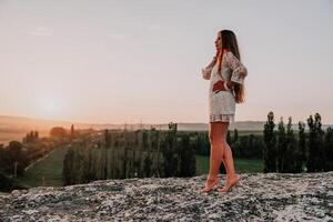 Happy woman in white boho dress on sunset in mountains. Romantic woman with long hair standing with her back on the sunset in nature in summer with open hands. Silhouette. Nature. Sunset. photo