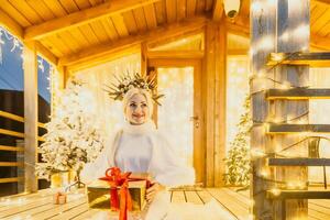 contento sonriente mujer en un blanco vestir participación un regalo caja en frente de un Navidad árbol. foto