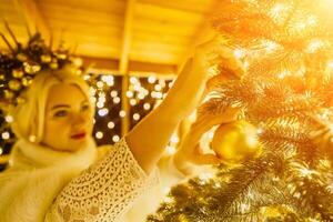 A blonde woman in white dress and a crown of gold ornaments decorate Christmas tree with gold ornaments and lights. The tree is decorated with gold balls and is lit up with lights. photo