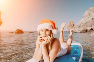 Woman sea sup. Close up portrait of happy young caucasian woman with long hair in Santa hat looking at camera and smiling. Cute woman portrait in a white bikini posing on sup board in the sea photo