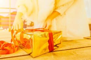 A woman in a white dress is holding a gold box with a red ribbon. She is wearing a crown on her head. The woman appears to be opening the gift box. photo