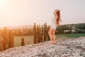 Happy woman in white boho dress on sunset in mountains. Romantic woman with long hair standing with her back on the sunset in nature in summer with open hands. Silhouette. Nature. Sunset. photo