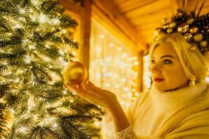 A blonde woman in white dress and a crown of gold ornaments decorate Christmas tree with gold ornaments and lights. The tree is decorated with gold balls and is lit up with lights. photo