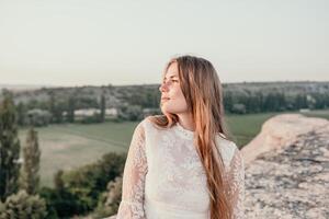 contento mujer en blanco boho vestir en puesta de sol en montañas. romántico mujer con largo pelo en pie con su espalda en el puesta de sol en naturaleza en verano con abierto manos. silueta. naturaleza. puesta de sol. foto