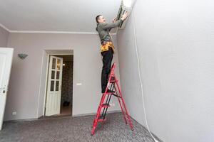 Professional electrician man with screwdriver maintaining, cleaning modern air conditioner indoors. Young technician standing on a ladder in the room repairing or installing air conditioner. photo