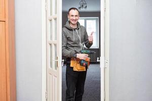 Maintenance Concept. Focused serious young locksmith with tattoo on hand wearing blue uniform standing on one knee in doorway installing doorknob handle, repairing wooden front door entrance photo