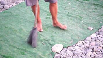 unrecognizable man sweeping the green carpet of artificial lawn on the beach from pebbles with broom. video