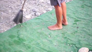 irreconocible hombre barriendo el verde alfombra de artificial césped en el playa desde guijarros con escoba. video