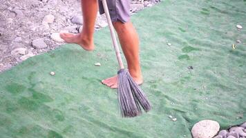 unrecognizable man sweeping the green carpet of artificial lawn on the beach from pebbles with broom. video