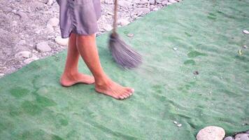 unrecognizable man sweeping the green carpet of artificial lawn on the beach from pebbles with broom. video