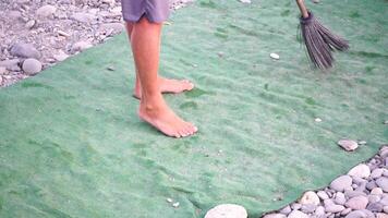 unrecognizable man sweeping the green carpet of artificial lawn on the beach from pebbles with broom. video