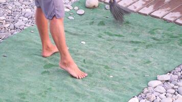 unrecognizable man sweeping the green carpet of artificial lawn on the beach from pebbles with broom. video