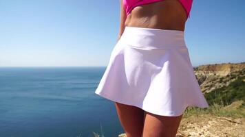 mujer verano viaje mar. contento rubia dama en un rosado camisa y blanco falda danza en mar Oceano acantilado. soleado día, capturar triunfo y admiración de asombroso vista, celebrando logro. video