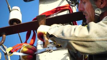 Skilled electrician in helmet fixes wires standing on ladder near high pole against blue sky on summer day backside view. Electrical service and mounting on the pole. Slow motion video