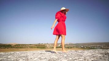 woman in a red dress and straw hat standing on a rock, enjoying the fresh air. Embracing freedom and beauty of nature. Happy lady on top of mountain with raised hands while wind is blowing her dress. video