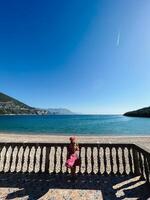 pequeño niña soportes cerca el barandilla en el playa y mira a el mar foto